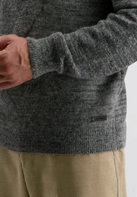 Grijze gebreide trui met een gewatteerd patroon, geribbelde boorden en zoom. Een klein zwart label is zichtbaar nabij de onderste rand.