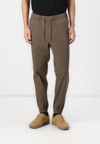 Osoba nosząca brązowe spodnie dresowe z regulowanym pasem i elastycznymi mankietami, w połączeniu z beżowymi sneakersami i czarnymi skarpetkami.