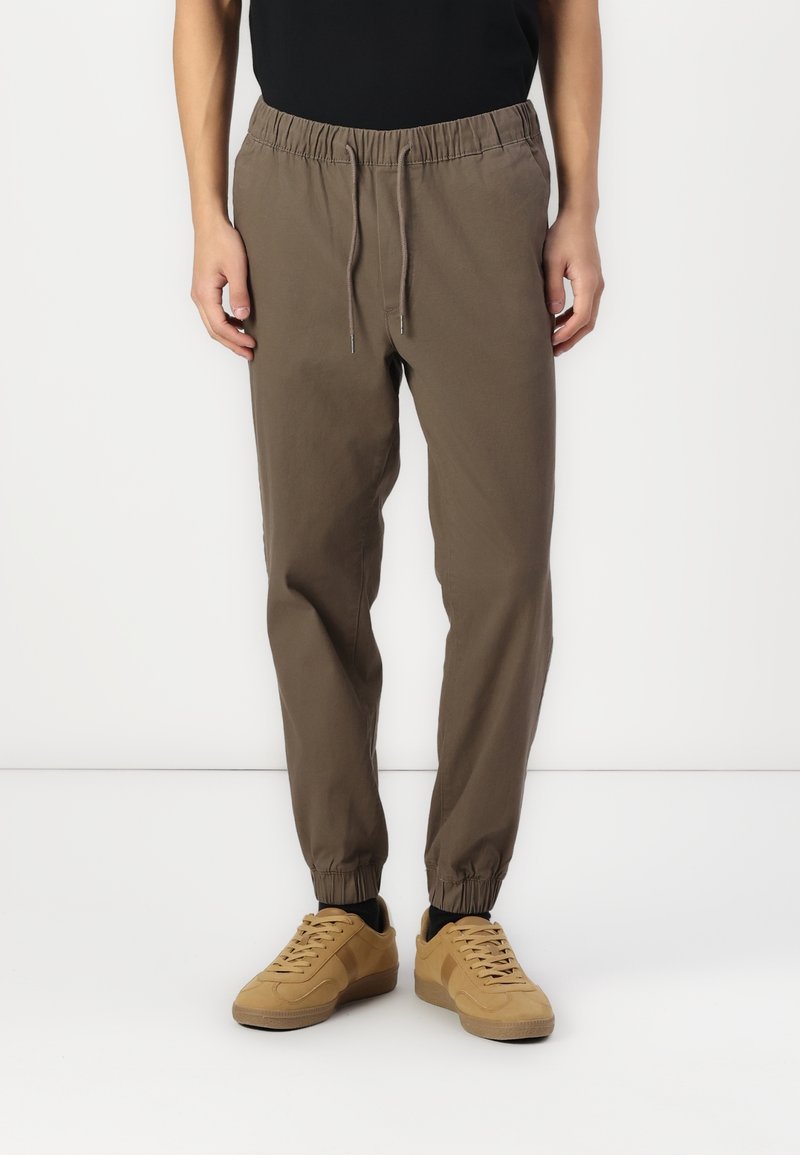 Osoba nosząca brązowe spodnie dresowe z regulowanym pasem i elastycznymi mankietami, w połączeniu z beżowymi sneakersami i czarnymi skarpetkami.