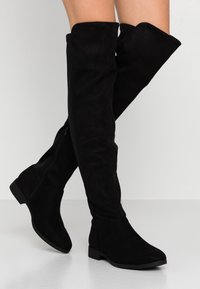 Bottes en daim noires montantes avec une semelle plate, dotées d'une fermeture éclair sur le côté et d'une texture lisse.