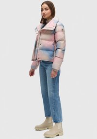 Giacca a vento imbottita in gradiente rosa e blu con colletto alto, zip metallica e bottoni a pressione. Abbinata a jeans azzurri chiari e stivaletti beige.