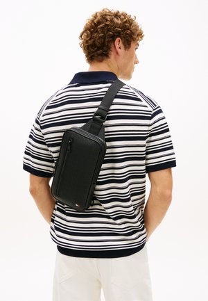 Młody mężczyzna z kręconymi włosami, noszący czarną torbę na ramię, w granatowo-białej koszulce w paski z krótkim rękawem i białych spodniach, zwrócony tyłem.
