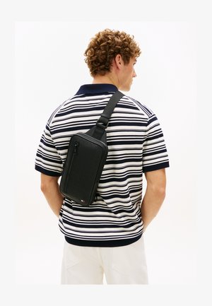 Jeune homme aux cheveux bouclés portant un sac bandoulière noir sur un t-shirt à manches courtes rayé bleu marine et blanc, ainsi qu'un pantalon blanc, vu de dos.