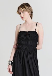 Junge Person mit kurzen blonden Haaren, die ein schwarzes, gerafftes Kleid mit dünnen Trägern und ein großes silbernes Armreif-Armband am linken Handgelenk trägt.
