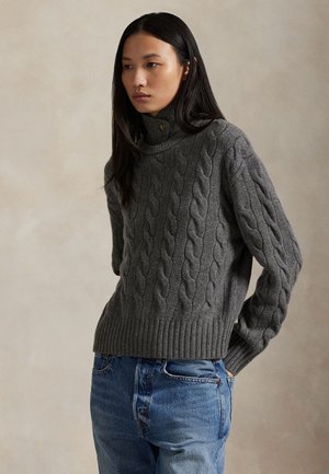 Femme aux longs cheveux foncés portant un pull à col roulé torsadé gris et un jean bleu, debout devant un fond neutre.