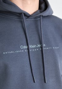 Capuchon sweatshirt in donkergrijze stof met een frontlogo dat leest "Calvin Klein Jeans" en "OPGERICHT NIGENTIG ACHT."