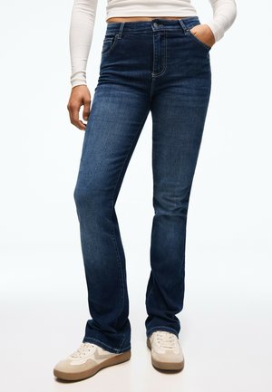 Personne portant un jean droit bleu foncé, un haut blanc à manches longues et des baskets beiges, une main dans la poche.