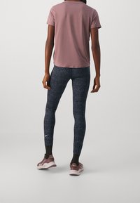 Osoba stojąca w różowej koszulce z krótkim rękawem, czarnych legginsach w wzory z białym logo Nike oraz w mauve'owych sneakersach Nike z czarnymi skarpetkami.