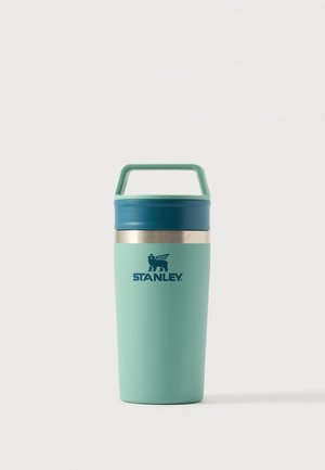 Mug de voyage isolant Stanley vert clair avec couvercle bleu sarcelle et poignée de transport sur un fond blanc uni.