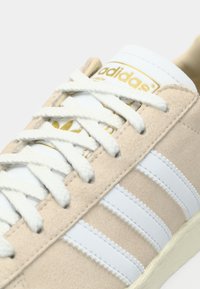 Bézs színű velúr sportcipő fehér bőr csíkokkal és fűzőkkel, arany Adidas logóval egy fehér nyelven. Texturált és sima felületek kontrasztja.