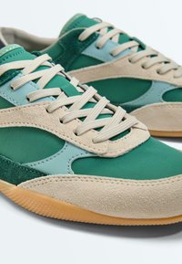 Baskets en daim vert et beige avec des accents bleu clair et des lacets beiges, dotées de semelles en caoutchouc naturel.