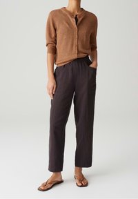 Osoba w brązowym swetrze zapinanym na guziki, luźnych czarnych spodniach z kieszeniami i brązowych klapkach, stojąca na jednolitym tle.