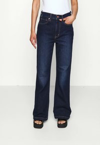 Mørkeblå vide jeans laget av denim, med kobbersting, frontknapping og fem lommer. Flate sko synlige.