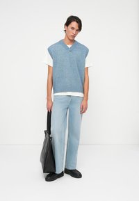 Colete de malha azul claro com decote em V, usado sobre uma t-shirt branca, combinado com jeans de ganga azul claro e sapatos pretos, segurando uma mala tote preta.