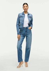 Γυναίκα που στέκεται φορώντας ανοιχτόχρωμο τζιν μπουφάν και τζιν, άσπρη μπλούζα και άσπρα ψηλά τακούνια, χαμογελώντας με το ένα χέρι στην τσέπη.