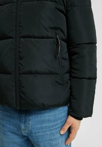 Chaqueta plumas negra con cierre frontal y bolsillo lateral. Presenta textura acolchada y puños acanalados. Combinada con unos vaqueros de mezclilla azules.