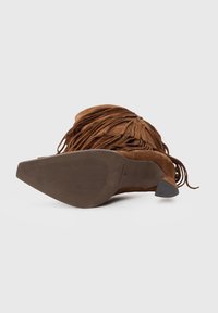 Stivale alla caviglia in suede marrone con dettagli in frange, punta appuntita e suola in gomma testurizzata, mostrato dal fondo.
