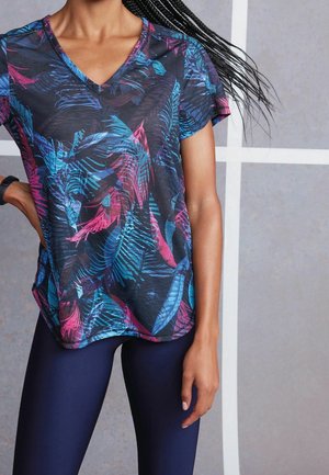 Woman wearing a V-neck, short-sleeve tee with blue and pink leaf patterns and dark fitted leggings, standing against a gray background.