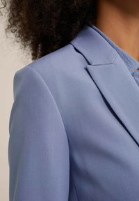 Jasnoniebieska marynarka o gładkiej fakturze, z klapą z wycięciem i usztywnionymi ramionami. Tkanina ma delikatny połysk.
