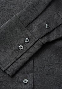 Camicia nera con texture e pieghe, dotata di un colletto con bottoni e bottoni grigi. Il tessuto appare leggero e morbido.