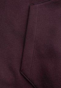 Material de hanorac burgund cu o textură netedă, având o mică buzunar înclinat pe față cu detalii de cusătură vizibile.