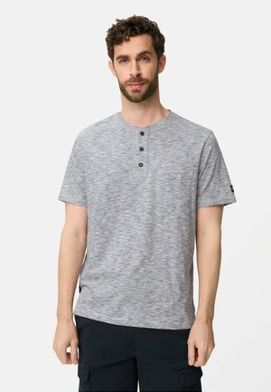 Mand med kort krøllet hår og skæg iført en lysegrå henley t-shirt med korte ærmer og tre knapper samt sorte bukser.