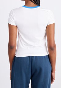 Bílé tričko s krátkým rukávem a modrým výstřihem, přiléhavý střih a hladká textura. Pohled zezadu s tmavě modrými kalhotami.