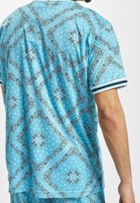 Hombre que lleva una camisa de manga corta color azul claro con patrones florales y geométricos en azul oscuro y blanco, mostrando la espalda y el brazo derecho.