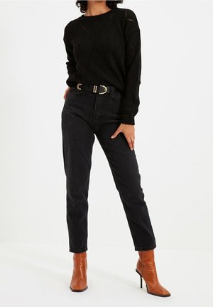 Personne portant un pull en maille noir, un jean noir taille haute avec une ceinture noire, et des bottines marron à talons, debout sur un fond blanc.