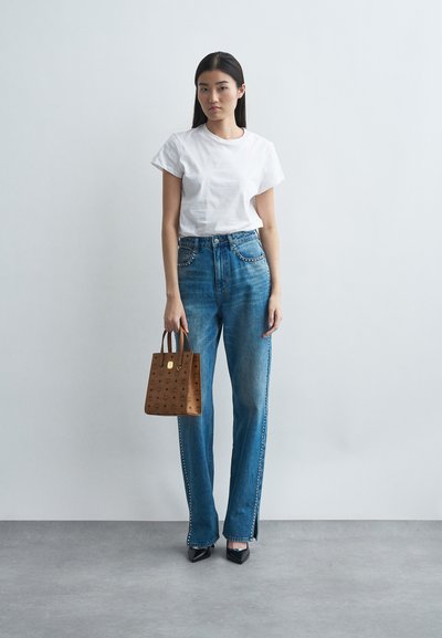 Nainen, joka käyttää valkoista t-paitaa, korkeavyötäröisiä sinisiä farkkuja, joissa on niititetyt sivusaumat, mustia kärkitossuja, ja hän pitää kädessään ruskeaa kuviollista laukkua.