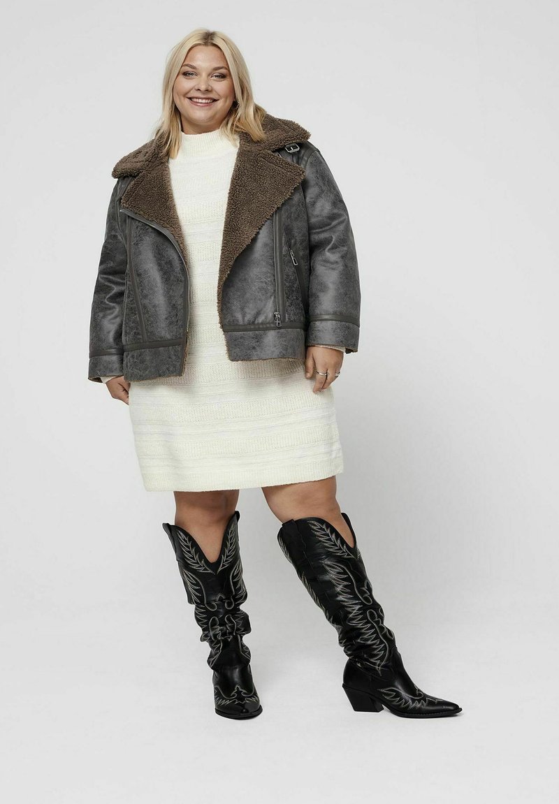 Chaqueta gris de gran tamaño con cuello de borrego marrón, vestido blanco texturizado y botas vaqueras negras hasta la rodilla con costuras decorativas.