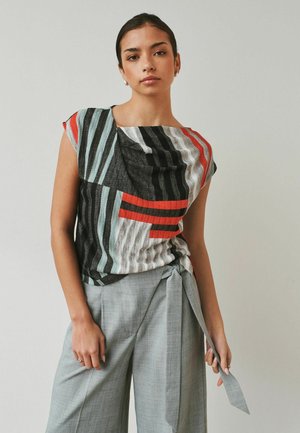 Woman with dark hair tied back wearing a multicolored striped top and high-waisted gray pants with a tied belt, standing against a plain background.