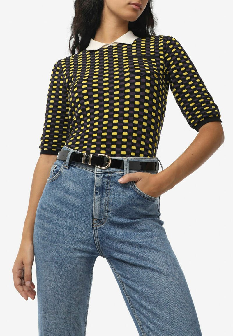 Top nero aderente con quadretti gialli e colletto bianco, abbinato a jeans blu con vita alta, sottolineato da una cintura nera.