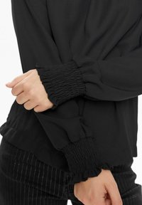 Top nero a maniche lunghe con polsini arricciati. Tessuto morbido, vestibilità rilassata, dettagli arricciati ai polsini, abbinato a pantaloni a righe nella parte inferiore.