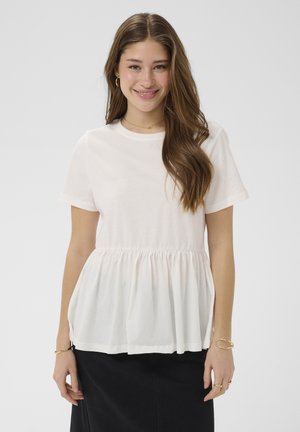 Młoda kobieta z długimi brązowymi włosami uśmiecha się, ubrana w biały krótki top peplum i czarną spódnicę na jednolitym tle.