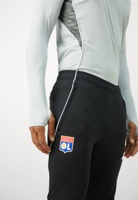 Camiseta deportiva de manga larga en gris claro con detalles en negro, combinada con pantalones negros que presentan un logo de Olympique Lyonnais en rojo y azul.