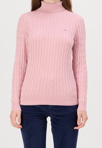 Pull à col roulé rose pâle avec texture côtelée et motif en torsades. Manches longues et coupe ajustée. Associé à un pantalon bleu marine foncé.