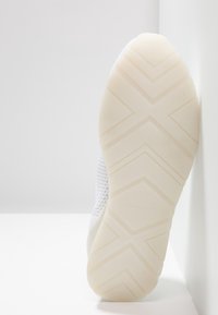 Baskets blanches avec une tige en mesh texturé, un bout arrondi, et une semelle en caoutchouc rainurée présentant un motif en zigzag et un logo en bas.