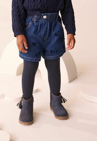 Mørkeblå denimshorts med en samlet talje og sløjfe-applikationer, parret med ribbede, mørke strømpebukser og blå ankelstøvler med frynser.