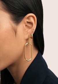 Pendientes de metal dorado en forma ovalada alargada con una barra vertical. Múltiples pequeñas piedras similares a diamantes en la parte superior de la oreja. Textura suave.