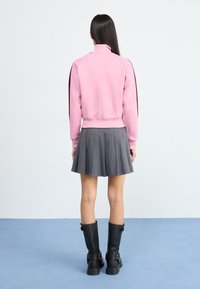 En person med langt mørkt hår iført en lyserød lynlås sweatshirt og en grå plisseret nederdel står på et lysblåt tæppe foran en neutral baggrund.