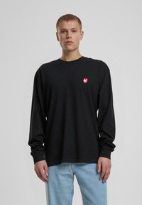 Schwarzes langärmliges T-Shirt aus Baumwolle mit einem kleinen roten Herzlogo auf der Brust, kombiniert mit hellblauen Jeans.