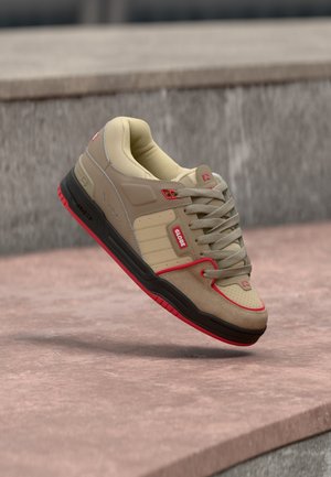 Beige en tan skateschoen met rode accenten en zwarte zool, met "GLOBE" logo, zwevend boven een rozeige betonnen ondergrond.