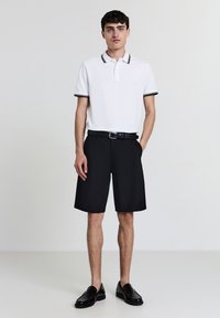 Polo bianco con bordi neri, abbinato a pantaloni corti neri. Il modello indossa una cintura nera e scarpe eleganti nere, in piedi su uno sfondo neutro.