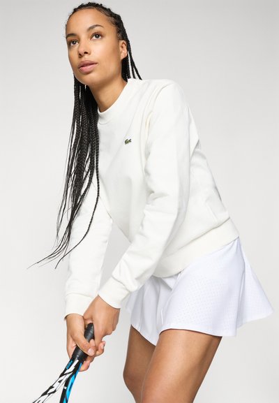 Sweatshirt blanc à col rond, à manches longues et portant un petit logo vert. Associé à une jupe de tennis blanche perforée, tenant une raquette.