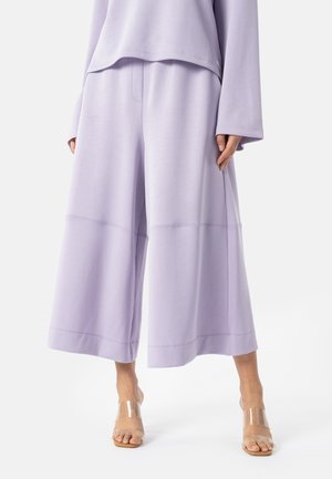 Kobieta ubrana w liliowe, szerokie spodnie o długości do połowy łydki, do tego dopasowany długi rękaw i przezroczyste sandały na obcasie, stojąca na białym tle.
