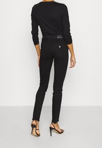 Černé skinny džíny s vysokým pasem, elastickým materiálem a trojúhelníkovým kovovým logem na zadní kapse. V kombinaci s černým topem s dlouhým rukávem a lodičkami.