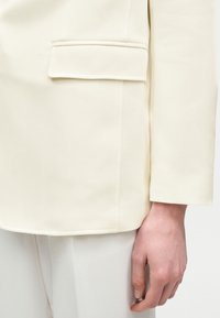 Gros plan sur la main d'une personne reposant à ses côtés, portant une veste de costume crème et un pantalon assorti.