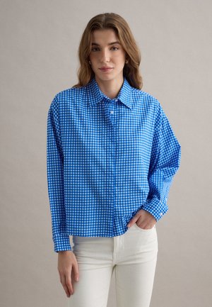 Junge Frau trägt ein blau-weiß kariertes Hemd mit Knöpfen und weiße Hose, steht vor einem schlichten hellen Hintergrund.