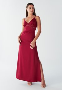 Bordo spalvos maksi suknelė plonomis petnešėlėmis, nėrinių akcentais prie liemens ir šoniniu skeltuku. Lygus, lengvai krintantis audinys su aptempta silueto linija.